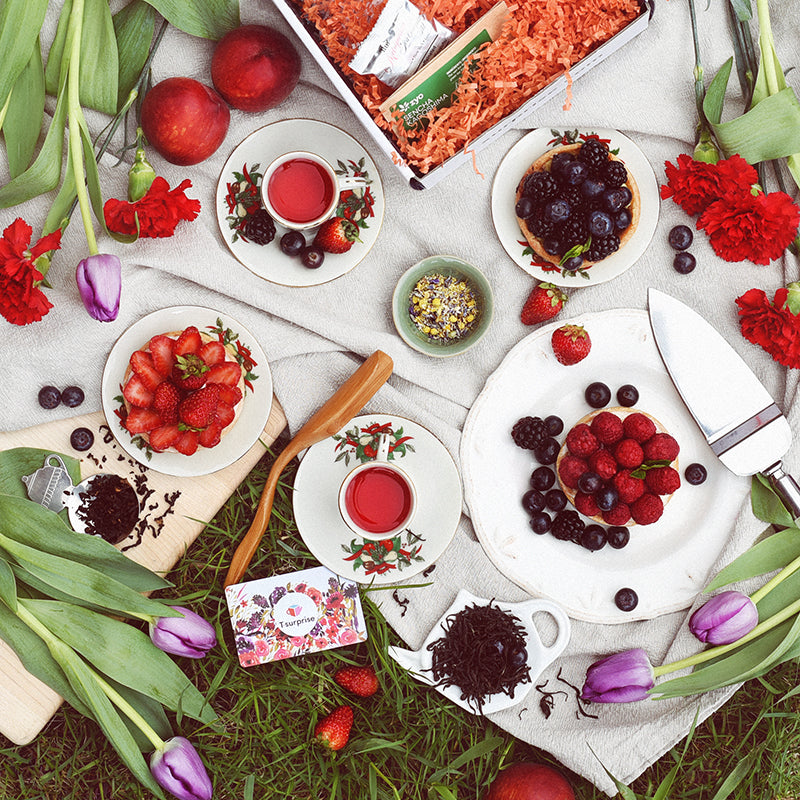 Table printanière avec tasse de thé rouge, tartelettes aux fraises et petits fruits, fleurs et herbe fraîche, thème Fleurs et lumière T•Surprise.